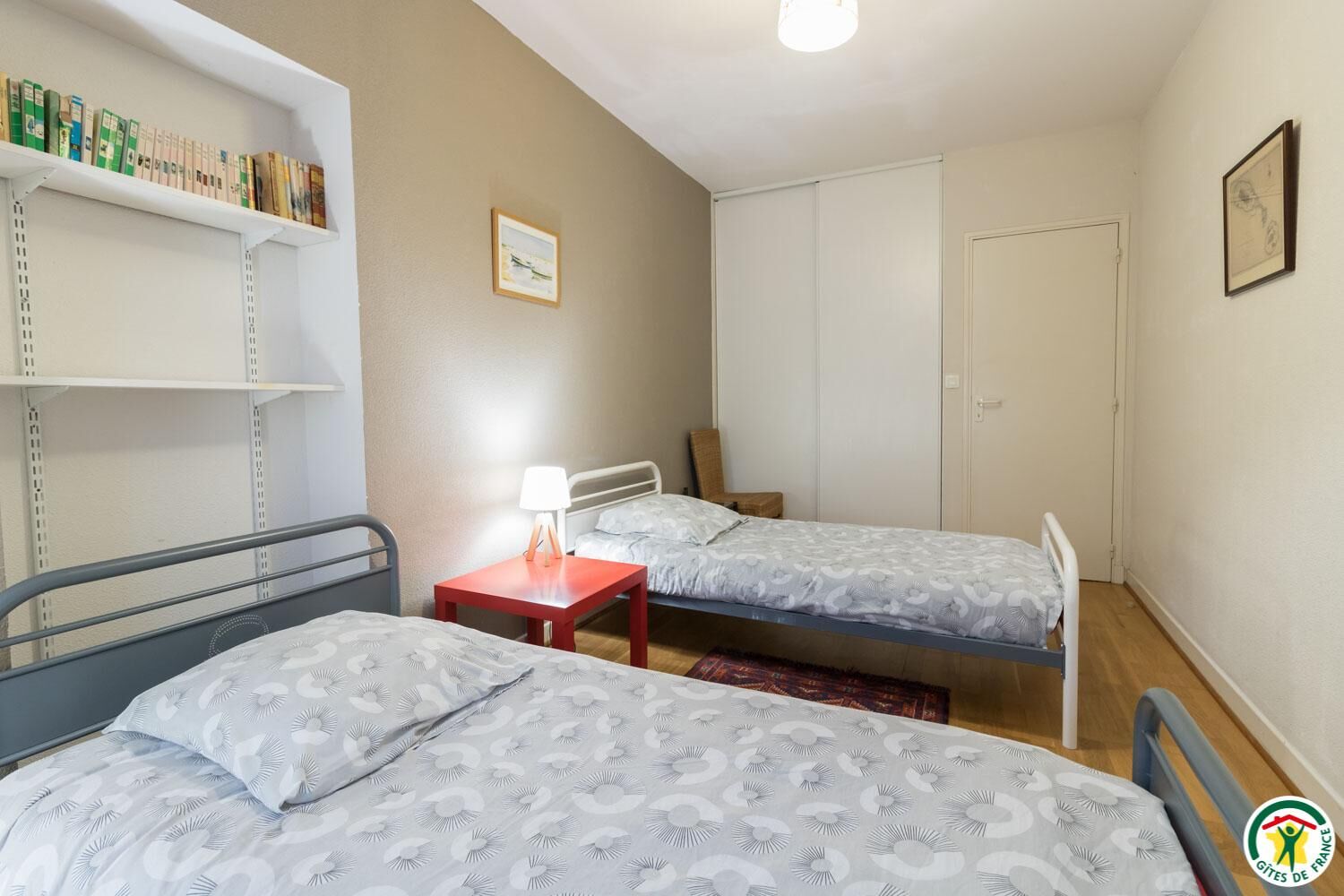 A small, tidy room with two single beds, a nightstand with a lamp, a bookshelf, and a door.