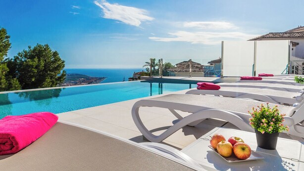 Un área junto a la piscina con sillones de descanso, una mesa con manzanas y una planta en maceta, y vista al mar y edificios al fondo.