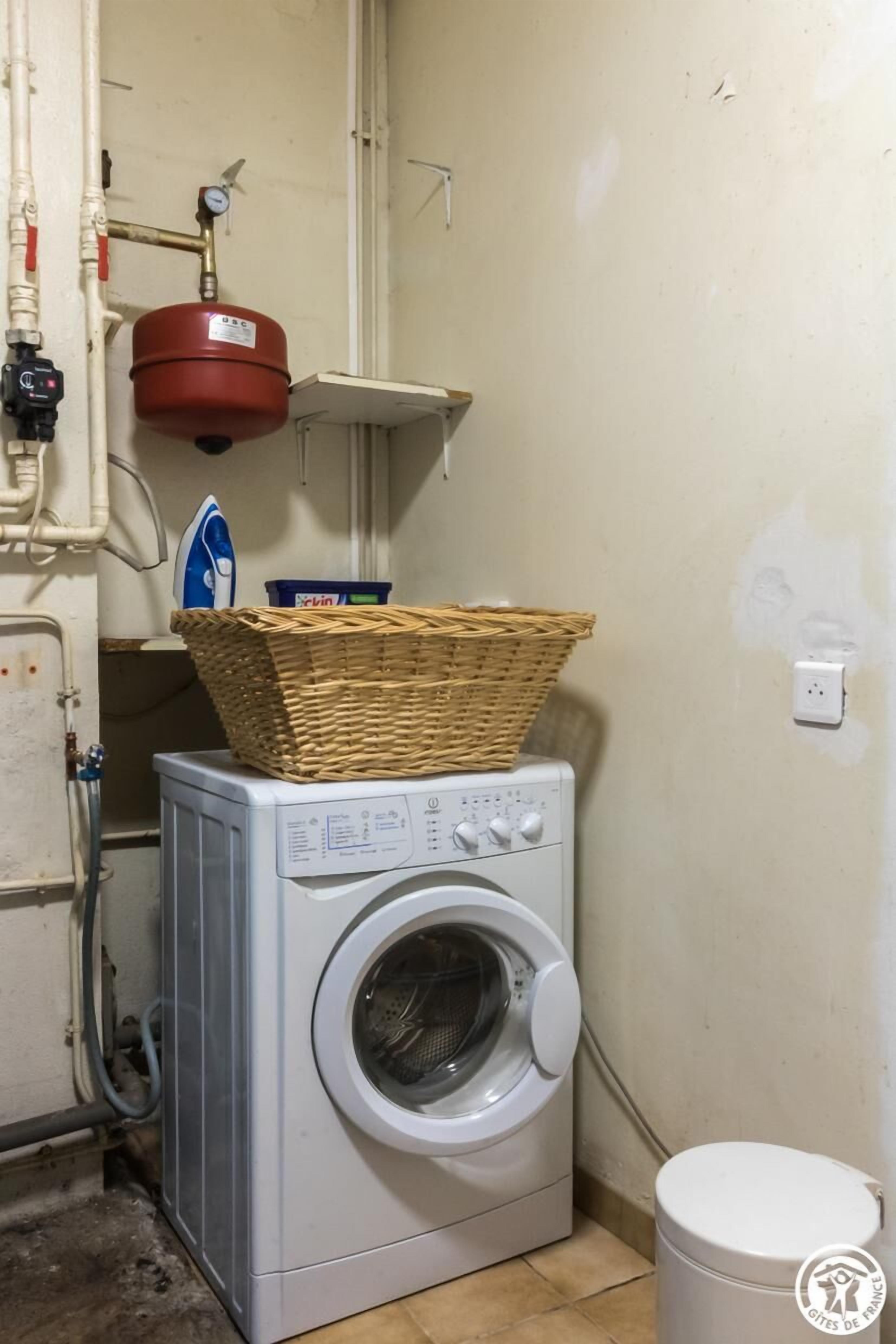 Ein kleiner Hauswirtschaftsraum mit Waschmaschine, rotem Durchlauferhitzer und einem Korb obenauf.