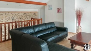 A living room with a black leather sofa, wooden ceiling beams, a stone wall, and a dining area with a wooden railing.