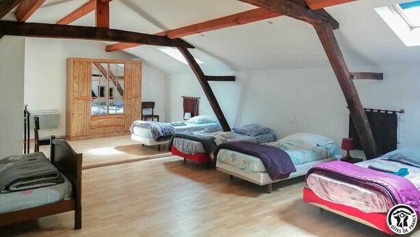 A room with a sloped ceiling, wooden beams, and a skylight. There are three beds with different bedding and a wooden wardrobe.