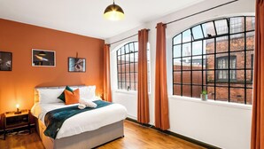 A hotel room with a large bed, a nightstand, a hanging light, and a view of a brick building through large windows. - 3 Bed Flat - Sleeps 7 - Parking - Wifi (Birmingham)
