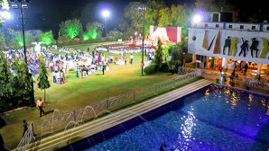 A large outdoor pool with a bridge leading to a grassy area with people and a stage. - Arristo Resorts & Club (Ahmedabad)