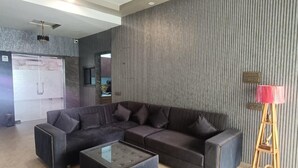 A modern living room with a sectional sofa, ottomans, and a glass coffee table. - Arristo Resorts & Club (Ahmedabad)