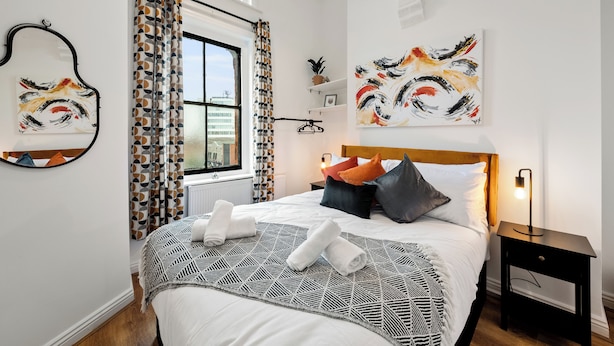 A neatly made bed with a geometric patterned bedspread, a bedside table with a lamp, and abstract paintings on the walls.