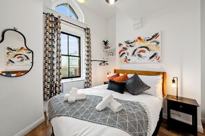 Ein ordentlich bezogenes Bett mit einem geometrisch gemusterten Überwurf, ein Nachttisch mit einer Lampe und abstrakte Gemälde an den Wänden.