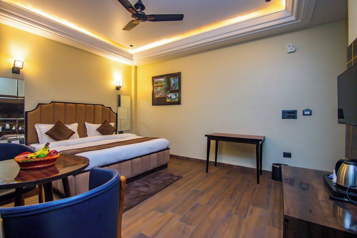 A hotel room with a bed, a desk, a chair, a small table, a ceiling fan, and a wall-mounted TV.