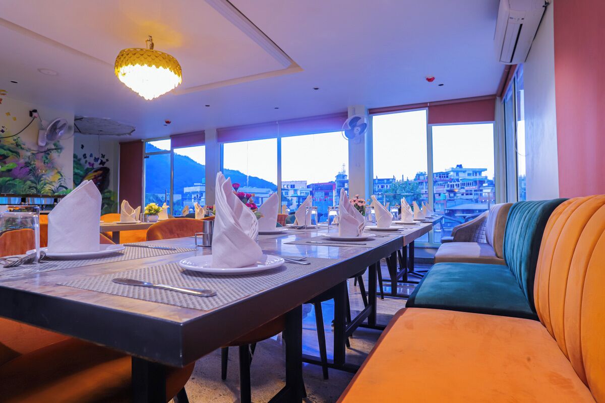 Un restaurante con una larga mesa dispuesta para comer, un sofá con cojines y grandes ventanales que ofrecen vistas a la ciudad.