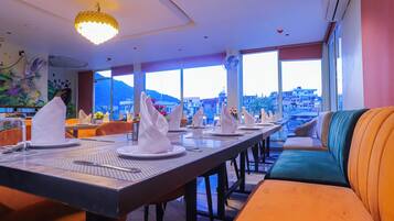 A restaurant with a long table set for dining, a sofa with cushions, and large windows offering a city view.