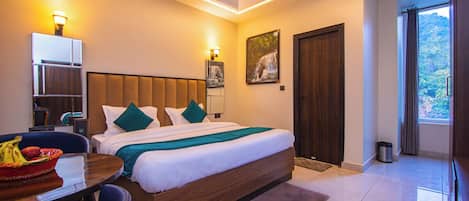 Habitación de hotel con una cama grande, un escritorio con una silla, una mesita con fruta, un cuadro en la pared y vistas a zonas verdes por la ventana.