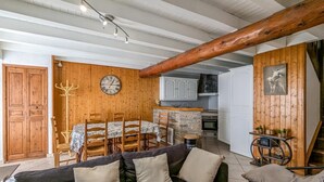 Ein gemütliches Wohnzimmer mit Holzbalkendecke, einem Esstisch mit Stühlen, einer schwarzen Couch mit Kissen und einer Küchenzeile mit Schränken und Herd.