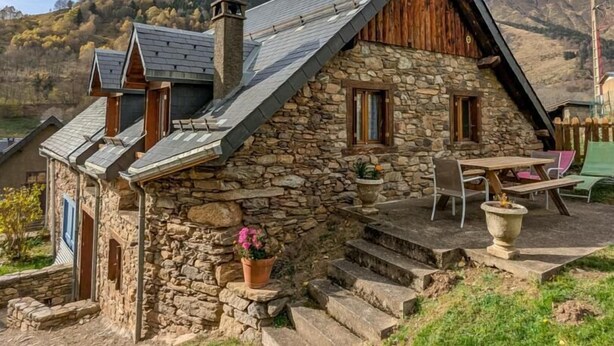 Ein Steinhaus mit geneigtem Dach, einer Satellitenschüssel und einer Terrasse mit Gartenmöbeln.