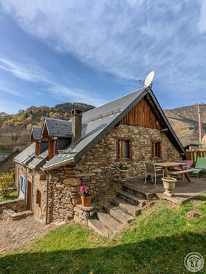 Une maison en pierre avec un toit en pente, une antenne parabolique et une terrasse aménagée avec du mobilier d’extérieur.