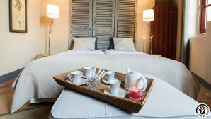 A neatly made bed with a tray of breakfast items, including cups, a teapot, and a candle, in a room with wooden shutters and a framed picture on the wall.