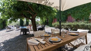 An outdoor dining area with a table set for two, surrounded by lounge chairs and a large umbrella providing shade.