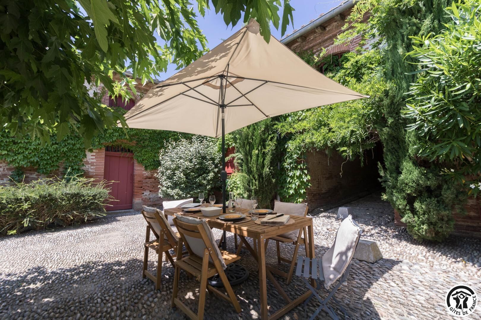 An outdoor dining area with a table and chairs under a large umbrella, surrounded by greenery and a brick wall.