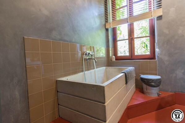 A bathroom with a bathtub, a window with blinds, and a trash bin.