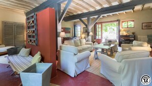 A spacious living room with a high ceiling and exposed beams, featuring a sofa set, a dining table, and a piano.
