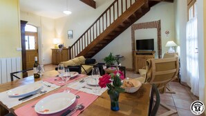 A dining area with a wooden table set for four, a staircase, a television, and a sofa in the background.