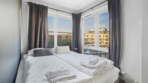 A double bed with white linens, two pillows, and a rolled towel. The room has a large window with curtains, a view of buildings outside, and a white ceiling.