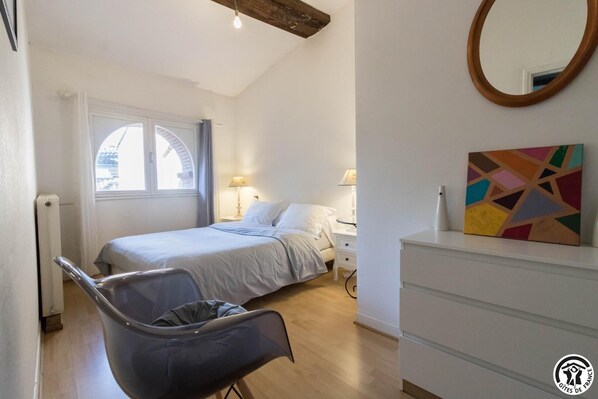 Un dormitorio con cama, mesitas de noche, una silla, cómoda, ventana con cortinas y un cuadro.