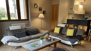 A living room with a grey sectional sofa, a coffee table, and a small ottoman. There is a radiator, a floor lamp, and a kitchen area in the background. - Gîte des Cyprès - Bérat - Gîte (Bérat)