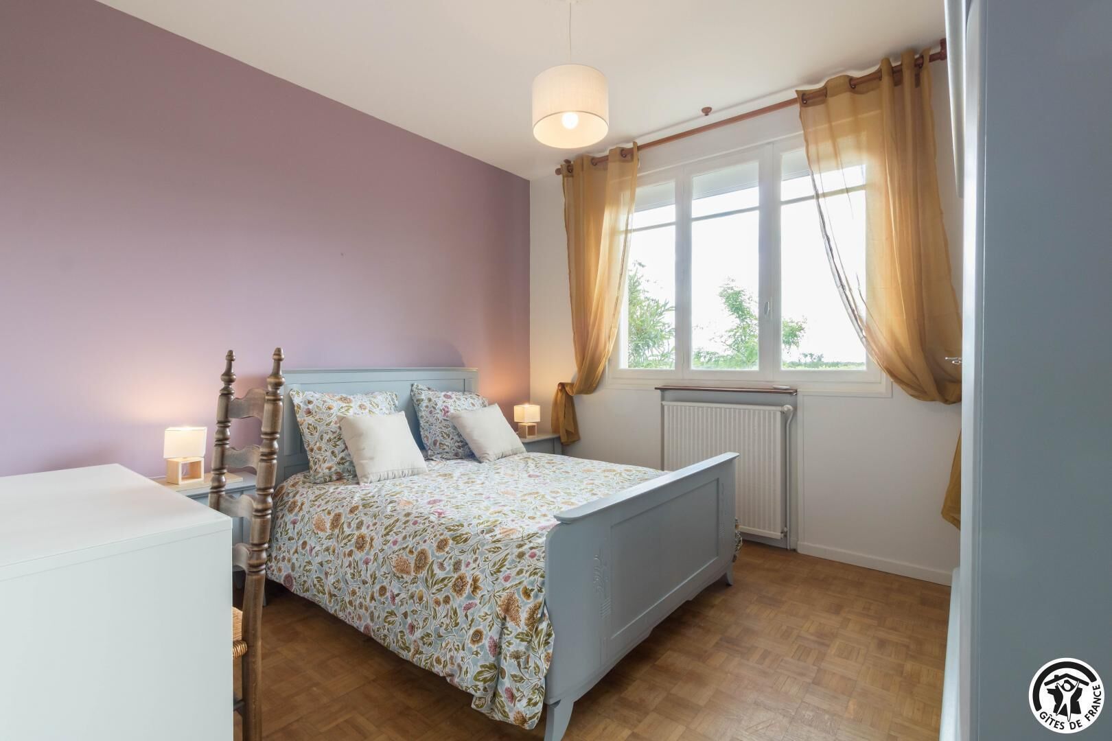 A bedroom with a bed, bedside lamps, a window with curtains, and a radiator.