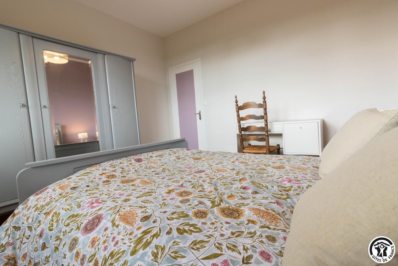 A bedroom with a floral bedspread, a wardrobe, a wooden chair, and a desk.