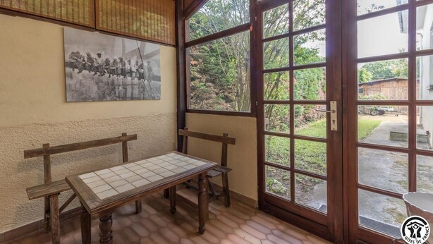 Una habitación con un banco de madera, una mesa de baldosas, un ventanal grande y un cuadro colgado en la pared.