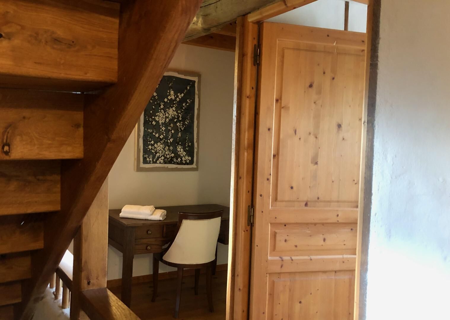 Un escalier en bois mène à une pièce dotée d’une porte en bois, d’une petite table avec une chaise et d’une décoration murale à motifs floraux.