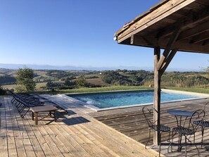 Una terraza de madera con piscina, mobiliario exterior y vistas a colinas ondulantes.