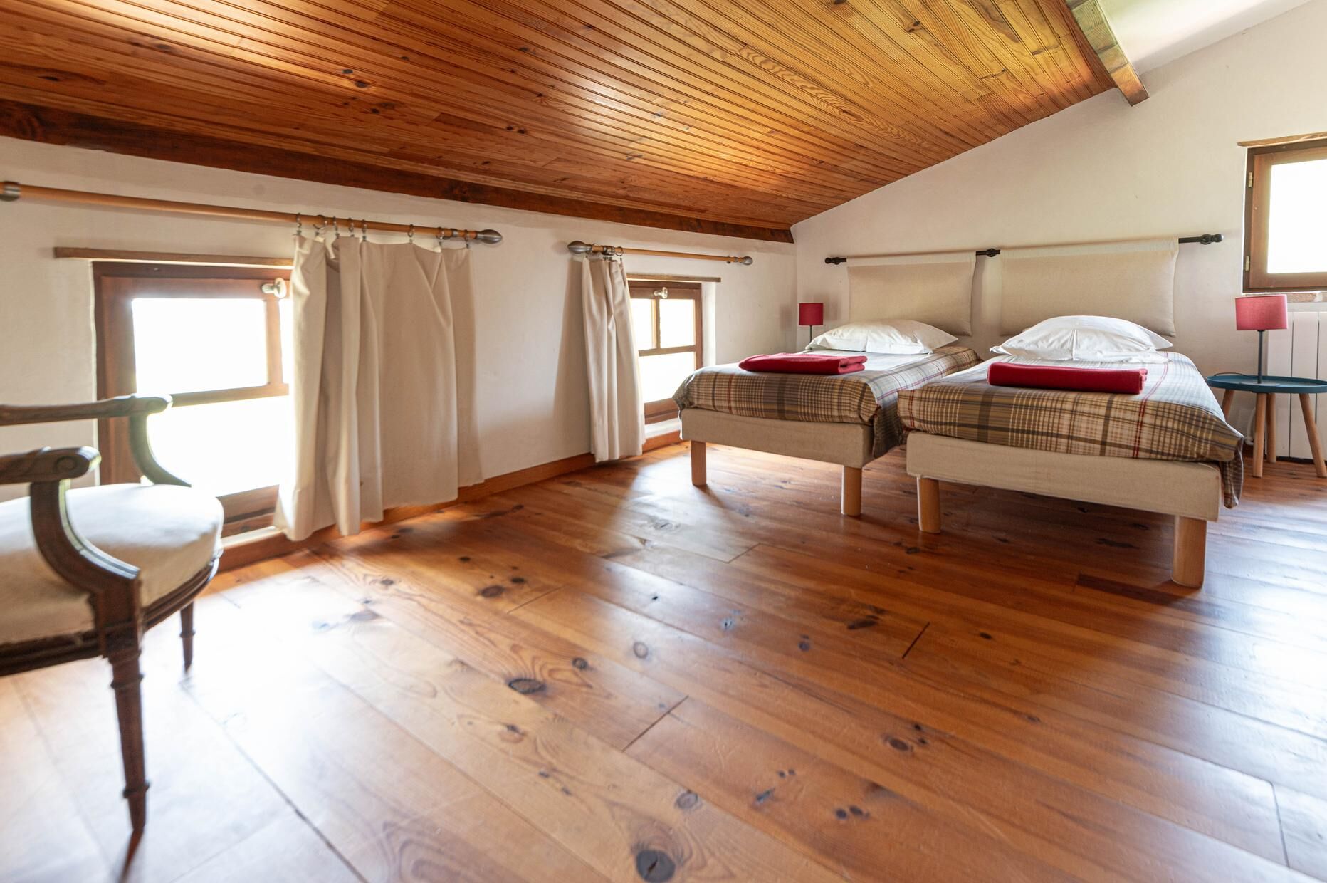 A room with two beds, wooden flooring, a chair, and a window with curtains.
