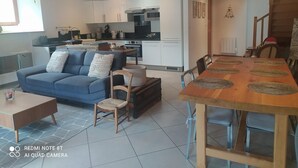 A modern living room with a sofa, coffee table, and dining table. A kitchen with white cabinets and appliances is visible in the background.