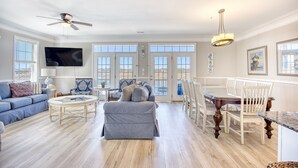 A spacious living room with a ceiling fan, a television, a grey sofa, and a dining area with wooden chairs and a table. - Cayman Villas Unit D-1 (North Myrtle Beach)