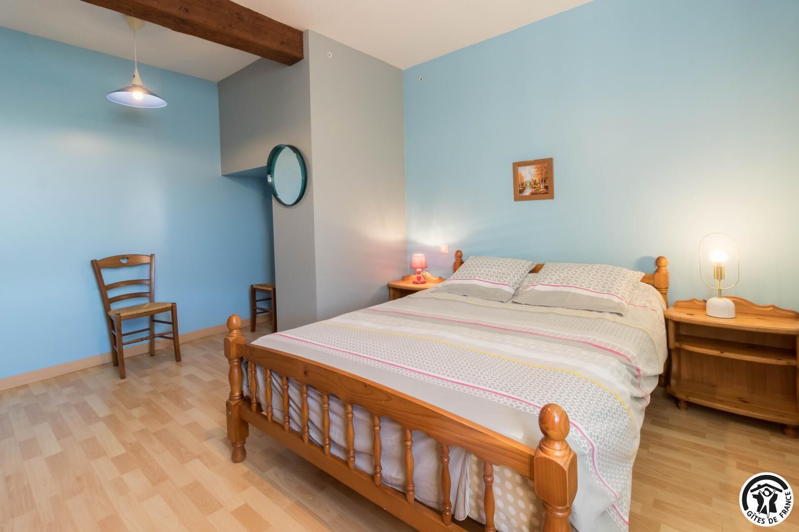 Une chambre à coucher avec un lit en bois, une chaise, une table de chevet avec une lampe et un miroir fixé au mur.