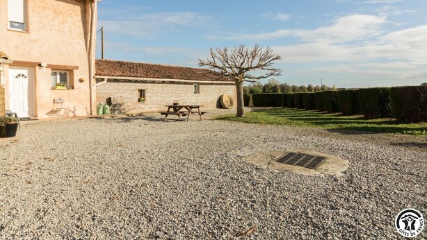 Un bâtiment rustique avec une allée de gravier, une table de pique-nique et un arbre au centre.