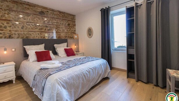 Un dormitorio con pared de piedra, una cama con ropa de cama blanca y roja, una mesita de noche y un ventanal con cortinas.