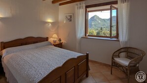 Une chambre à coucher avec un lit en bois, une table de chevet avec une lampe, un fauteuil en osier et une fenêtre donnant sur les montagnes.