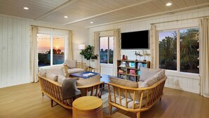 Ein gemütliches Wohnzimmer mit Holzboden, einer Couch, einem Couchtisch und einem Bücherregal. Große Fenster mit Vorhängen, ein an der Wand befestigter Flachbildfernseher und Blick auf den Sonnenuntergang.