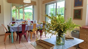 Un coin repas avec une table dressée pour quatre personnes, entourée de chaises, d’un vase de fleurs et d’une fenêtre donnant sur un paysage verdoyant.