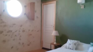 A bedroom with a wooden ceiling, a round window, a bed with white bedding, and a nightstand with a lamp. - Les Coteaux - Riolas (Riolas)