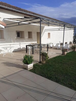Un patio exterior con pérgola, sillas blancas y una mesa.