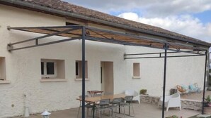 A covered patio area with a table and chairs, potted plants, and a white building with a tiled roof. - Les Coteaux - Riolas (Riolas)