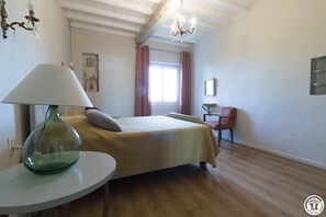 Un dormitorio con cama, una silla, una mesa con lámpara y una ventana con cortinas.