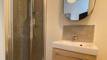 Een badkamer met een douchecabine van glas, een wit wastafeltje met een spiegel erboven en houten kastjes.