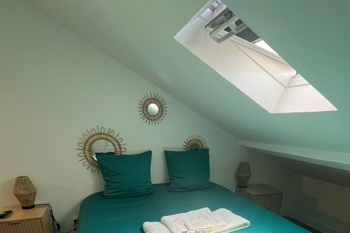 A bedroom with a green bedspread, two pillows, a bedside table with a lamp, and a skylight above the bed.
