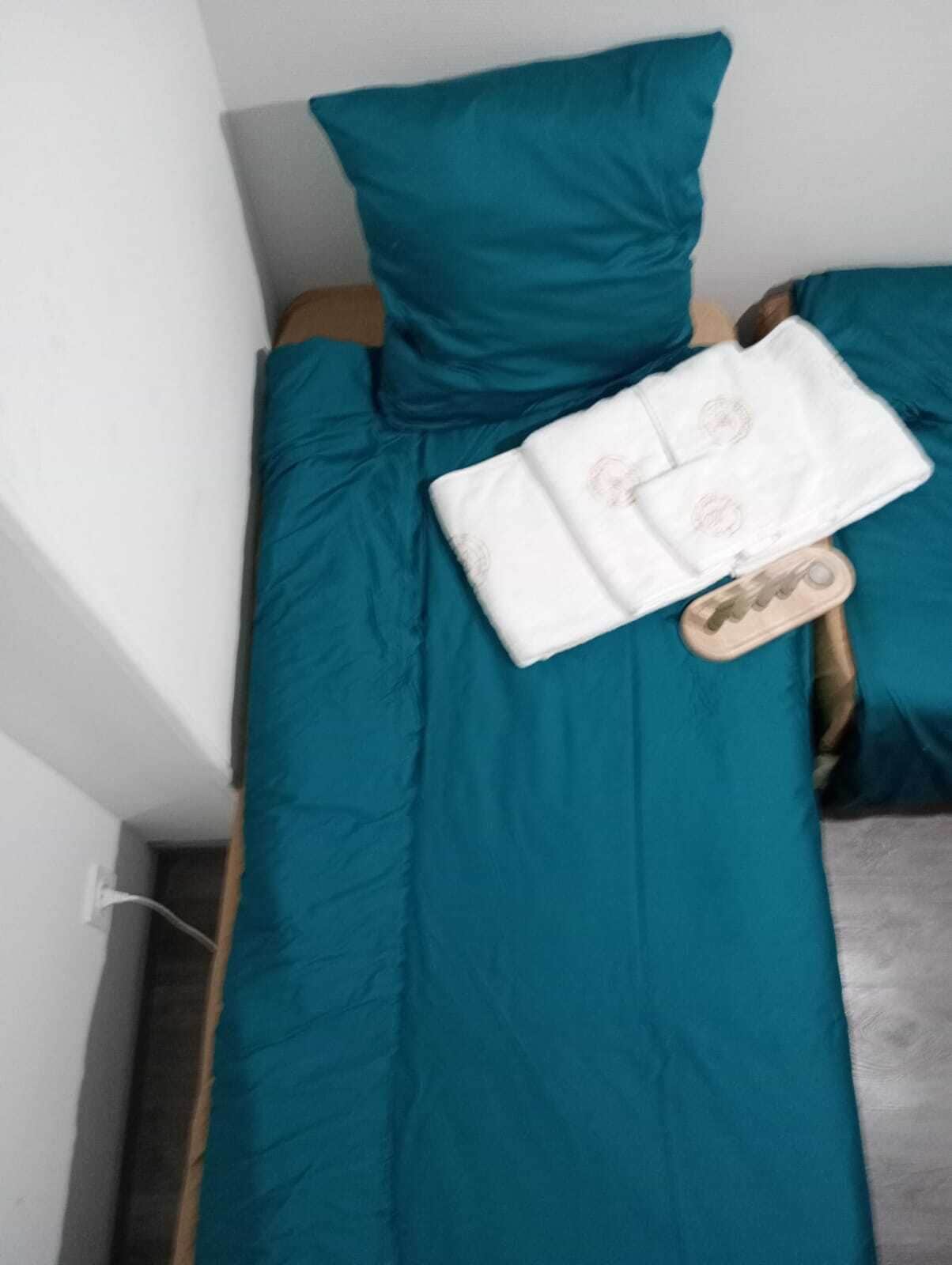 A single bed with teal bedding, a white pillow, and a folded white towel with a circular logo.