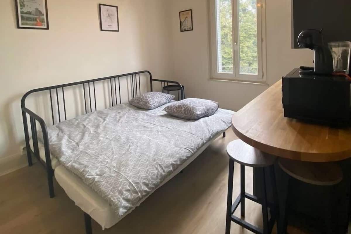 A small, single-bed room with a wooden table and a chair, a window with a view of greenery, and a ceiling light fixture.