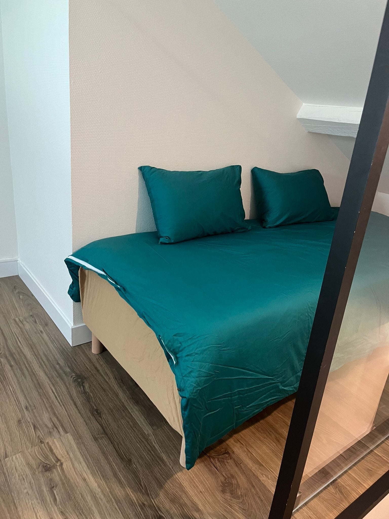 A bed with teal bedding in a room with wooden flooring and a textured beige wall.