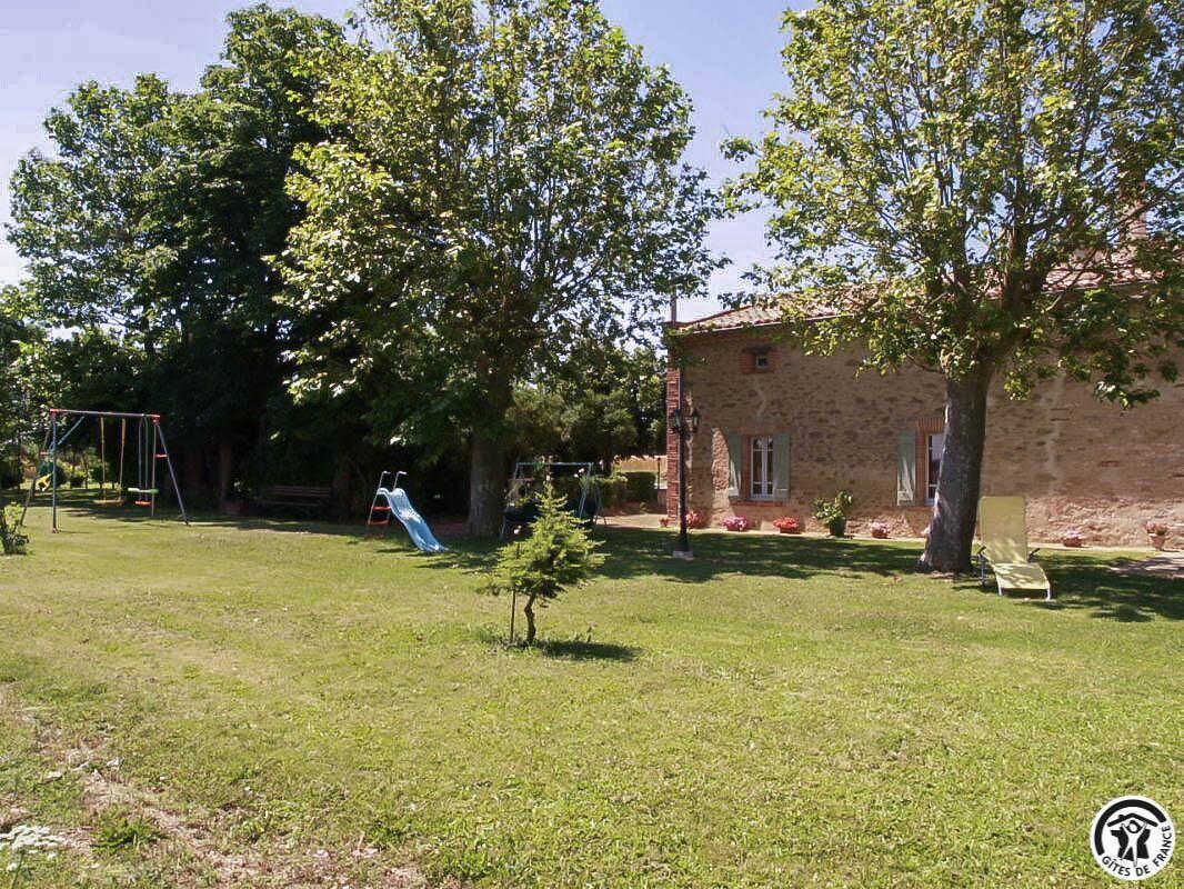 A grassy yard with a swing set, a small tree, and a stone building with a red roof.